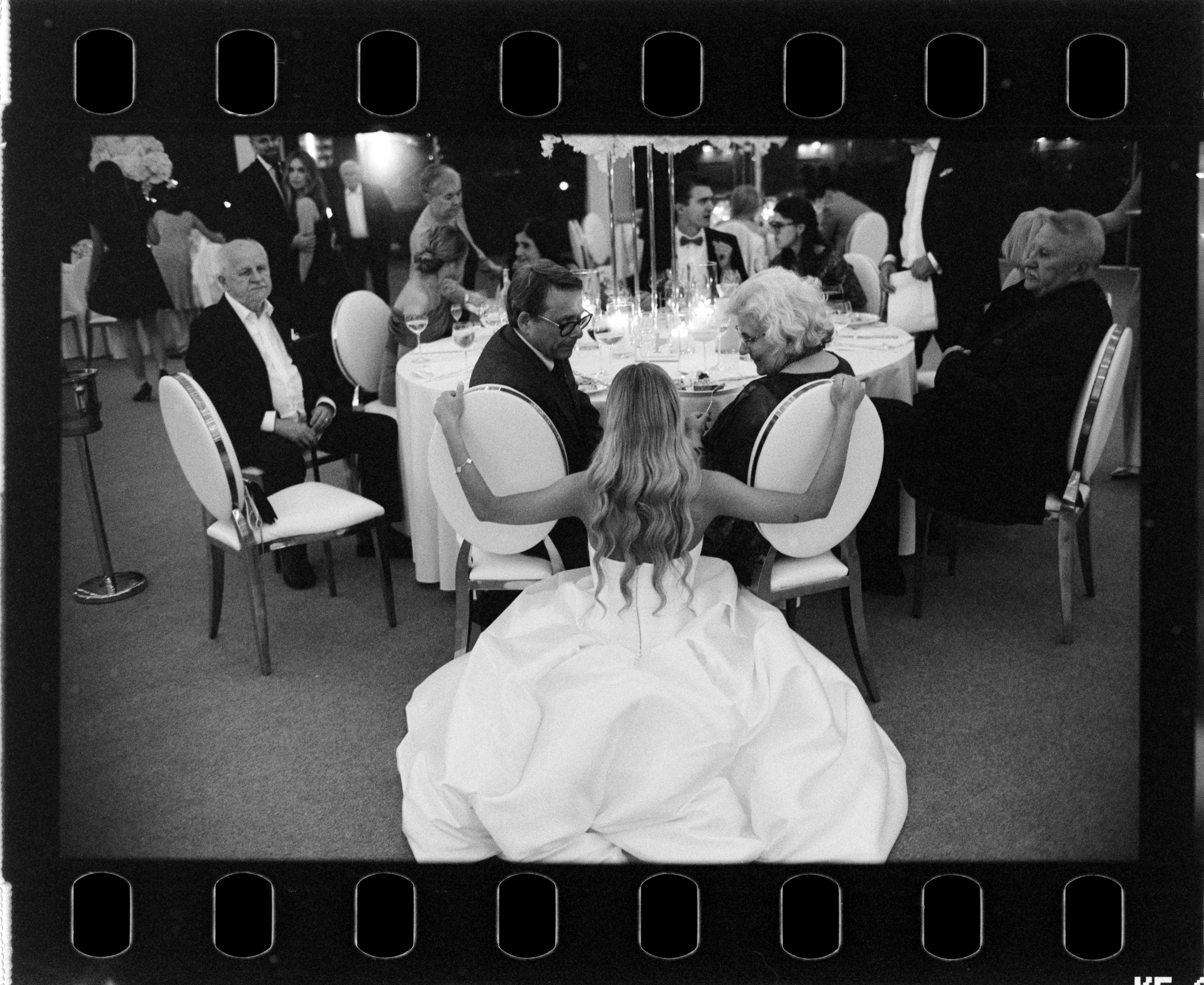 Panna młoda siedzi przy stole z bliskimi, widok od tyłu. Fotografia na filmie Ilford HP5.