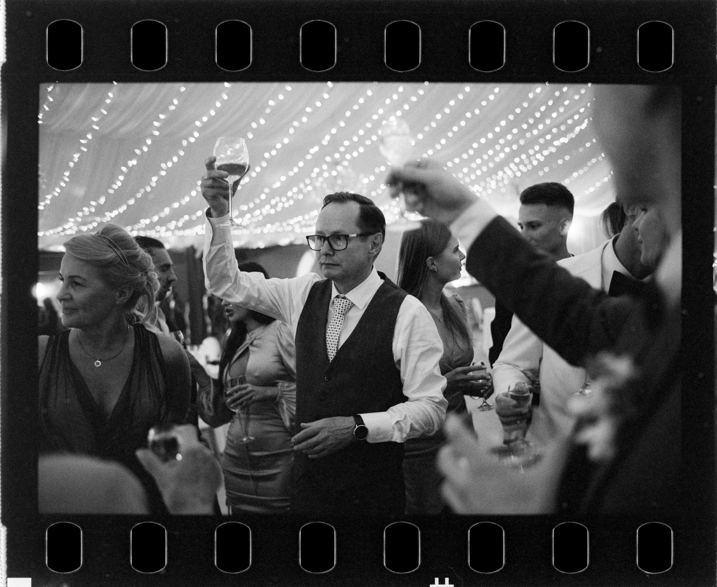 Goście wznoszą kieliszki podczas toastu. Fotografia na filmie Ilford HP5.