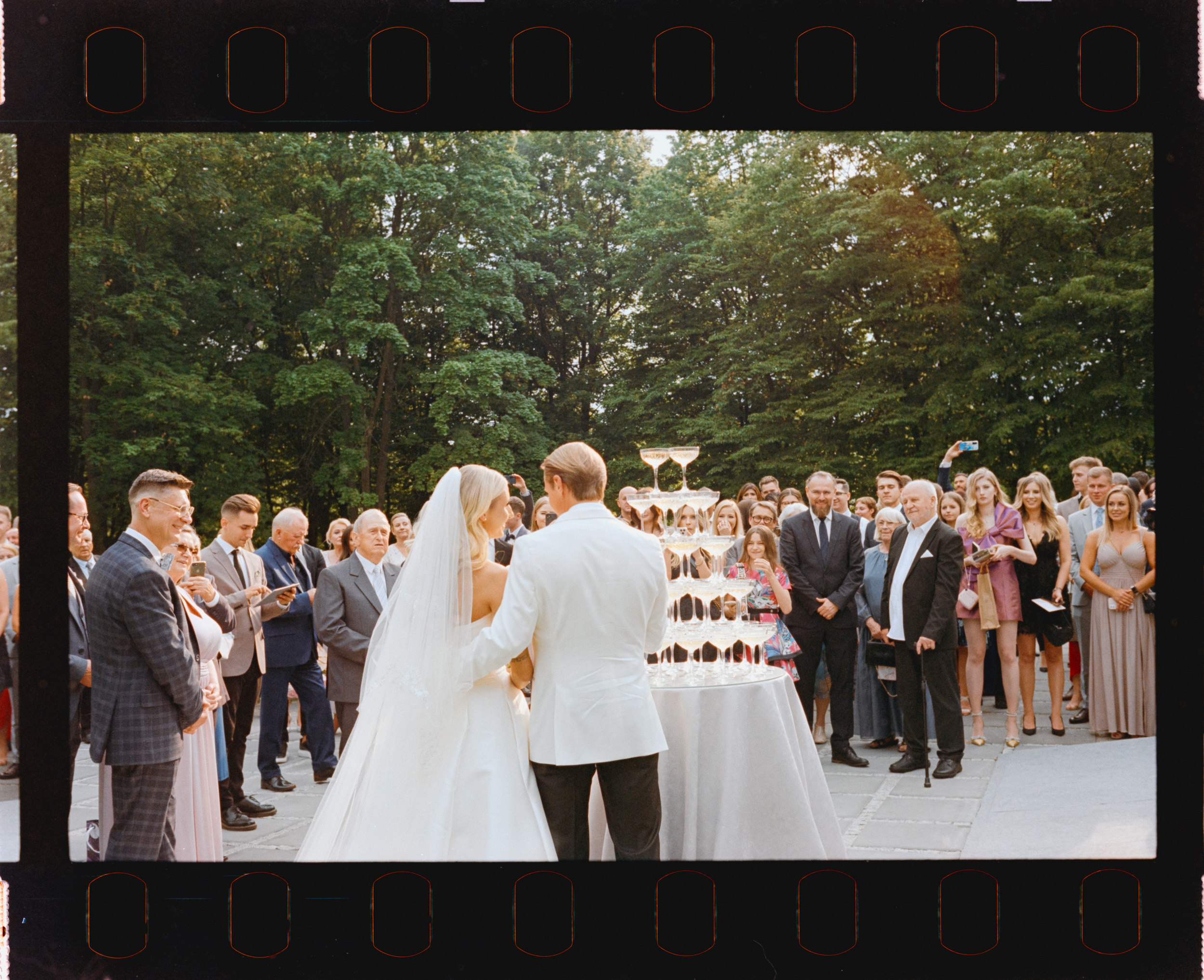Nowożeńcy przy piramidzie kieliszków w otoczeniu gości. Fotografia na filmie Kodak UltraMax.